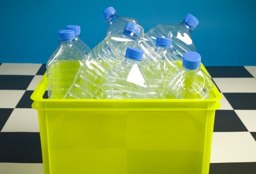 A bright lime green plastic container filled with several transparent plastic bottles with blue caps, some of which are slightly crushed or leaning. The bottles appear to be used for beverages and are empty, with visible ridges along their sides. The container is situated on a black and white checkered surface, resembling a tiled floor, with a solid blue wall in the background. This setup suggests a typical indoor environment for waste collection or disposal, relevant to rubbish removal services in West Ham. The overall scene is well-lit with even lighting, emphasizing the clarity of the plastic bottles and the vibrant color of the container, illustrating an example of waste or recycling collection commonly handled by Business Waste Removal West Ham in the local area