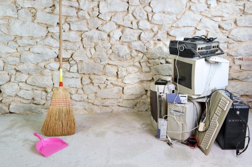 In a room with a textured light-colored stone wall in the background, a group of discarded electronic appliances and cables are stacked in the right foreground, including an old computer monitor, various desktop towers, and a tangled assortment of cords and peripherals. To the left, a small yellow-handled broom with natural bristles leans against the wall, while a pink plastic dustpan lies on the light-colored concrete floor, slightly tilted. The scene appears to be an indoor storage or disposal area, possibly associated with waste removal services in West Ham, with the focus on unwanted electronic waste and cleaning tools gathered in one corner. The lighting is natural and even, highlighting both the texture of the wall and the different materials of the discarded items, reflecting typical rubbish collection or clearance efforts carried out by companies like Business Waste Removal West Ham.