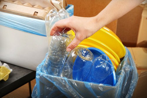 A close-up view of a person's hand gripping a clear plastic water bottle, positioned above a collection of recyclable plastic containers inside a large, partially open blue trash bag. The plastic containers include a yellow lid, a blue lid, and a blue container with a marked surface, indicating they are used for food or storage purposes. In the background, a white bin with a blue liner is visible on the left, sitting on a black surface, possibly a table or countertop, with part of a cardboard box and some crumpled paper nearby. The scene appears to be an indoor space, likely a kitchen or storage area, with warm lighting that emphasizes the textures of the plastics and the smoothness of the bottle. The overall setting is consistent with waste sorting or preparation for rubbish removal, aligned with services provided by Business Waste Removal West Ham, in the context of domestic or commercial rubbish collection in or around East London.